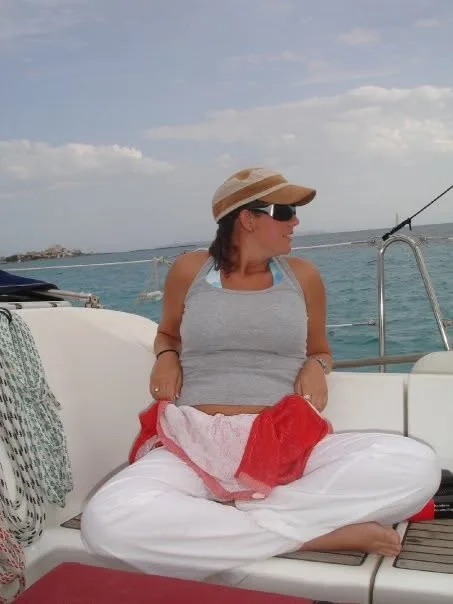 Pamela in a cap and sunglasses sitting cross-legged on a boat in Mallorca, turquoise sea behind her